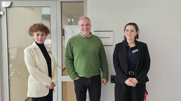 Nes und Anne-Marie beim Besuch der Kriminalpolizeidirektion Zwei Frauen und ein Mann stehen im Gang vor einer Türe mit dem Schild Staatsschutz
